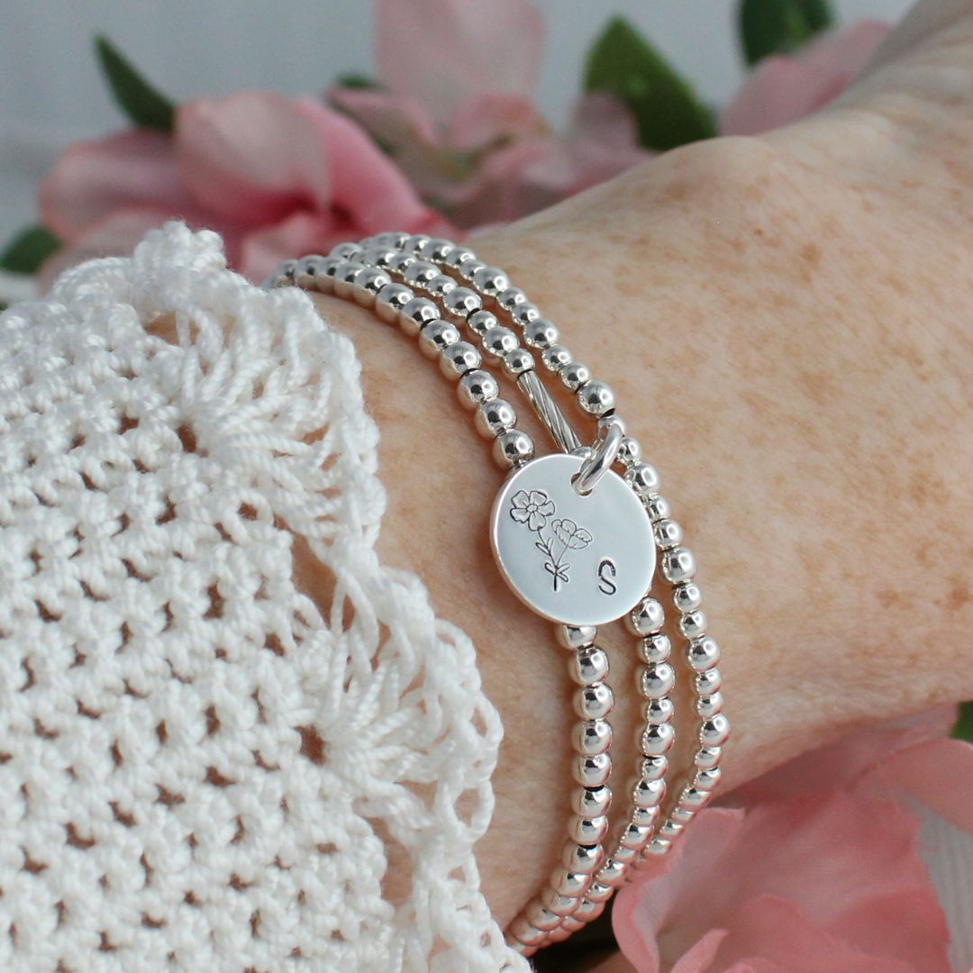 Silver beaded bracelet with a round charm on a wrist, against a blurred floral background.