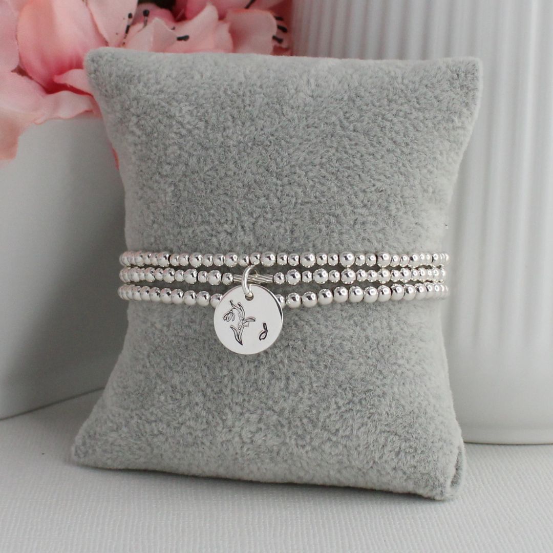 Silver bracelet with a round charm on a gray pillow with pink flowers in the background