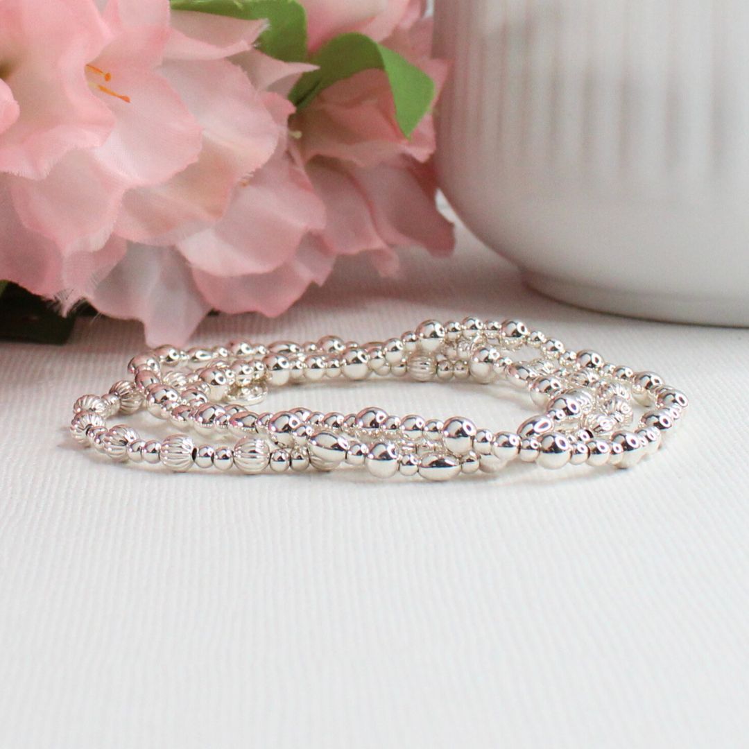 Silver beaded bracelet stack on a white surface with pink flowers in the background